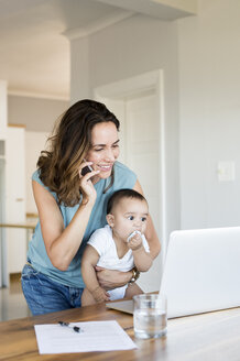 Mutter telefoniert, während sie einen kleinen Jungen trägt und auf einen Laptop schaut - CAVF39499