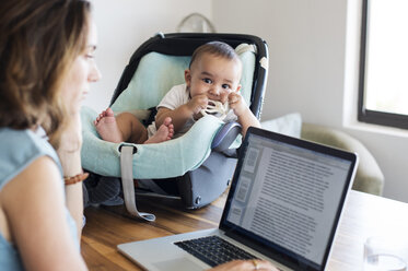 Mother using laptop computer by baby boy in seat on table at home - CAVF39504