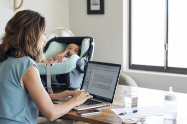 Mother using laptop computer by baby boy relaxing in seat on table at home - CAVF39505