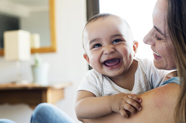 Happy mother looking at baby boy while sitting at home - CAVF39510