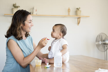 Mother feeding baby boy sitting on table at home - CAVF39518