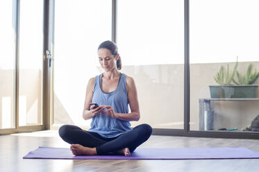 Woman using smart phone while sitting on exercise mat at home - CAVF39526