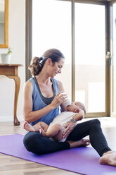 Smiling mother feeding son while sitting on exercise mat at home - CAVF39532