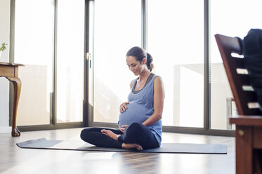 Schwangere Frau hält ihren Bauch, während sie zu Hause auf einer Übungsmatte sitzt - CAVF39534