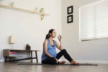 Schwangere Frau trinkt Wasser, während sie zu Hause auf einer Gymnastikmatte sitzt - CAVF39535