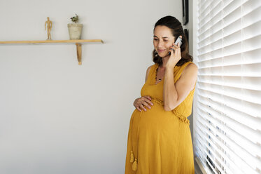 Schwangere Frau telefoniert mit ihrem Smartphone, während sie zu Hause am Fenster steht - CAVF39536