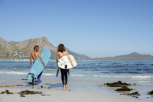 Rückansicht von Freunden, die ein Surfbrett tragen, während sie an einem sonnigen Tag am Strand spazieren gehen - CAVF39555