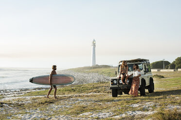 Man with surfboard walking towards female friends at beach against clear sky - CAVF39583