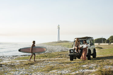 Mann mit Surfbrett geht auf Freundinnen am Strand zu gegen klaren Himmel - CAVF39583