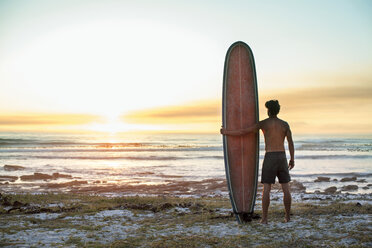 Rückansicht eines Mannes ohne Hemd, der ein Surfbrett am Strand vor einem klaren Himmel bei Sonnenuntergang hält - CAVF39604