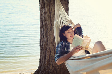 Frau liest ein Buch und entspannt sich in einer Hängematte unter einem Baum am Seeufer - CAVF39622