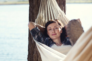 Portrait of woman relaxing on hammock by tree at lakeshore - CAVF39625