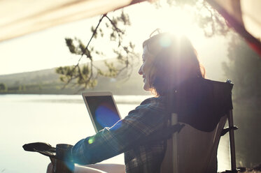 Lächelnde Frau mit Tablet-Computer entspannt sich auf einem Sessel am Seeufer an einem sonnigen Tag - CAVF39640
