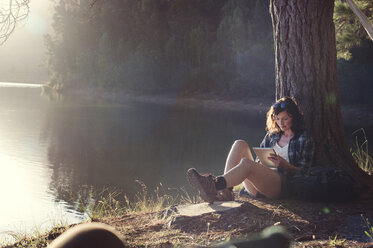 Woman using tablet computer while sitting by tree at lakeshore - CAVF39642