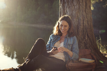 Smiling woman using mobile phone while relaxing at lakeshore - CAVF39644