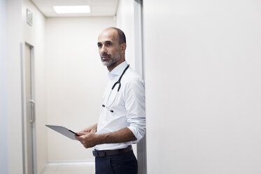 Arzt mit Tablet-Computer schaut weg, während er an einer Tür im Korridor steht - CAVF39657