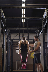 Männlicher Sportler, der einer Frau bei einer Übung an den Gymnastikringen im Fitnessstudio hilft - CAVF39680