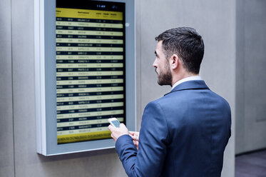 Rückansicht eines Geschäftsmannes, der sein Smartphone in der Hand hält, während er die Ankunfts- und Abfahrtstafel in einer U-Bahn-Station liest - CAVF39706