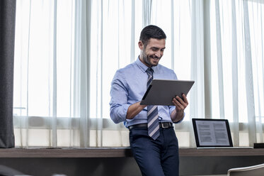 Happy businessman using tablet computer while standing by window in hotel room - CAVF39735