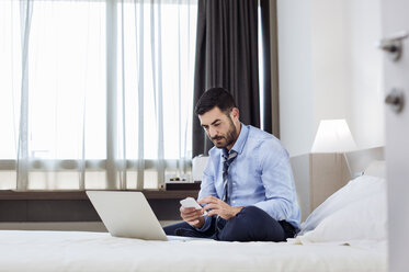 Geschäftsmann, der sein Smartphone benutzt, während er auf dem Bett im Hotelzimmer sitzt - CAVF39738