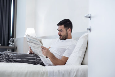 Man reading newspaper while relaxing on bed in hotel room - CAVF39742