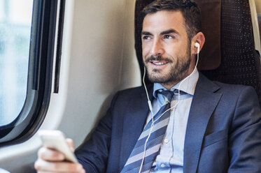 Smiling businessman listening music through smart phone in train - CAVF39750