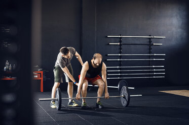 Trainer, der einem männlichen Athleten beim Heben einer Langhantel im Fitnessstudio hilft - CAVF39777