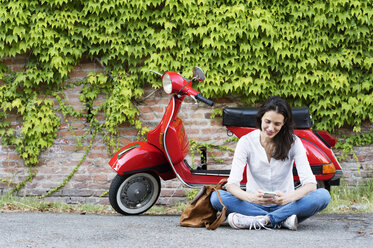 Frau, die ein Mobiltelefon benutzt, während sie an einem Motorroller und einer Efeuwand sitzt - CAVF39820