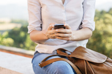 Mittelteil einer Frau, die ein Smartphone benutzt, während sie auf der Stützmauer einer Terrasse sitzt - CAVF39828