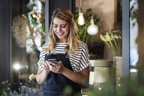 Florist, der ein Mobiltelefon benutzt, während er am Eingang eines Blumenladens steht - CAVF39852