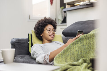 Smiling woman using phone while lying on sofa at home - CAVF39879