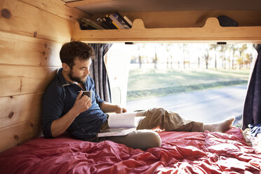 Man with mug reading book while sitting on bed in camper van - CAVF39928