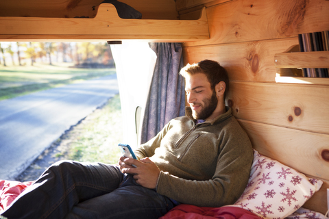 Mann benutzt Smartphone, während er auf dem Bett im Wohnmobil sitzt, lizenzfreies Stockfoto