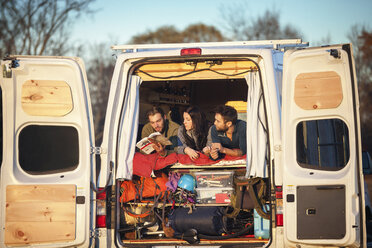 Couple looking at friend reading book while relaxing on bed in camper van - CAVF39936