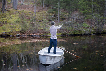 Rear view of man fishing while standing in boat on lake at forest - CAVF39939