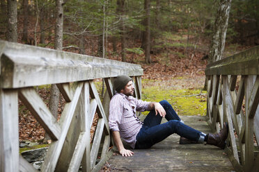 Side view of man sitting on footbridge in forest - CAVF39955