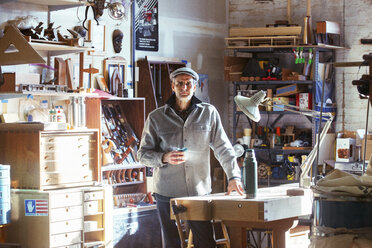 Porträt eines selbstbewussten Schreiners, der ein Getränk in der Hand hält, während er am Tisch in der Werkstatt steht - CAVF39967