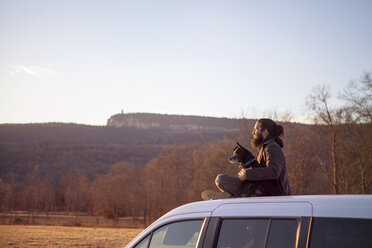 Seitenansicht eines Mannes, der mit seinem Hund auf dem Autodach sitzt, bei klarem Himmel - CAVF39999