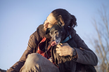 Zärtlicher Mann, der seinen Hund umarmt, während er vor einem klaren blauen Himmel sitzt - CAVF40000
