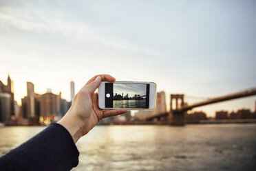 Cropped hand of woman photographing skyline and Brooklyn Bridge at dusk - CAVF40028