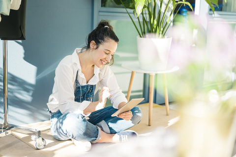 Lächelnde junge Freiberuflerin, die in ihrem Studio auf dem Boden sitzt und ein Tablet benutzt, lizenzfreies Stockfoto