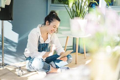 Lächelnde junge Freiberuflerin, die in ihrem Studio auf dem Boden sitzt und ein Tablet benutzt - MOEF01015