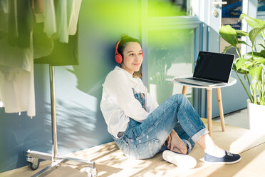 Smiling young freelancer sitting on the floor in her studio listening music with headphones and laptop - MOEF01017