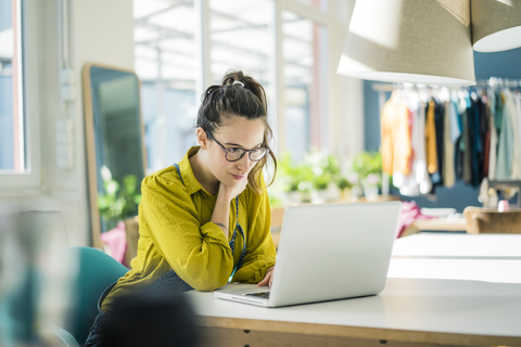 Modedesignerin sitzt am Schreibtisch in ihrem Atelier und schaut auf ihren Laptop, lizenzfreies Stockfoto