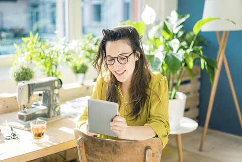 Porträt einer lächelnden Modedesignerin, die in ihrem Atelier sitzt und ein Tablet benutzt - MOEF01026