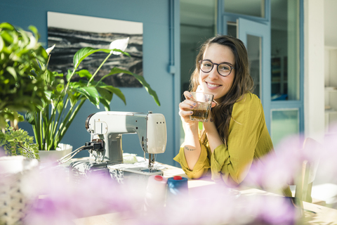 Porträt einer zufriedenen Modedesignerin, die mit einem Getränk am Schreibtisch in ihrem Atelier sitzt, lizenzfreies Stockfoto