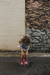 Verspieltes Mädchen springt in Pfütze auf Straße gegen Wand - CAVF40124