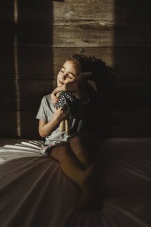 Girl with eyes closed embracing stuffed toy while sitting on bed against wood in darkroom - CAVF40157