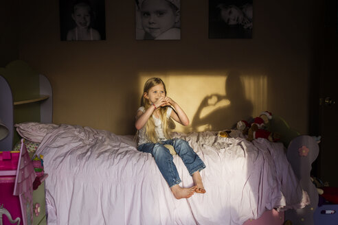 Girl making heart shape with hands while sitting on bed at home - CAVF40299