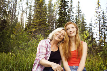 Thoughtful smiling mother and daughter sitting against trees at forest - CAVF40344